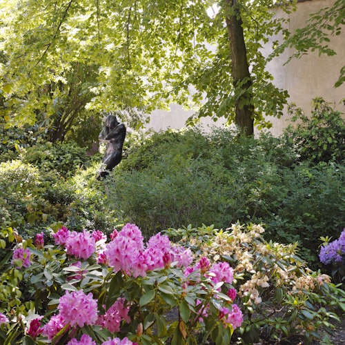Bujan vrt s jarkim ružičastim i žutim cvijećem, gustim zelenim lišćem i tamnim kipom djelomično skrivenim među drvećem.