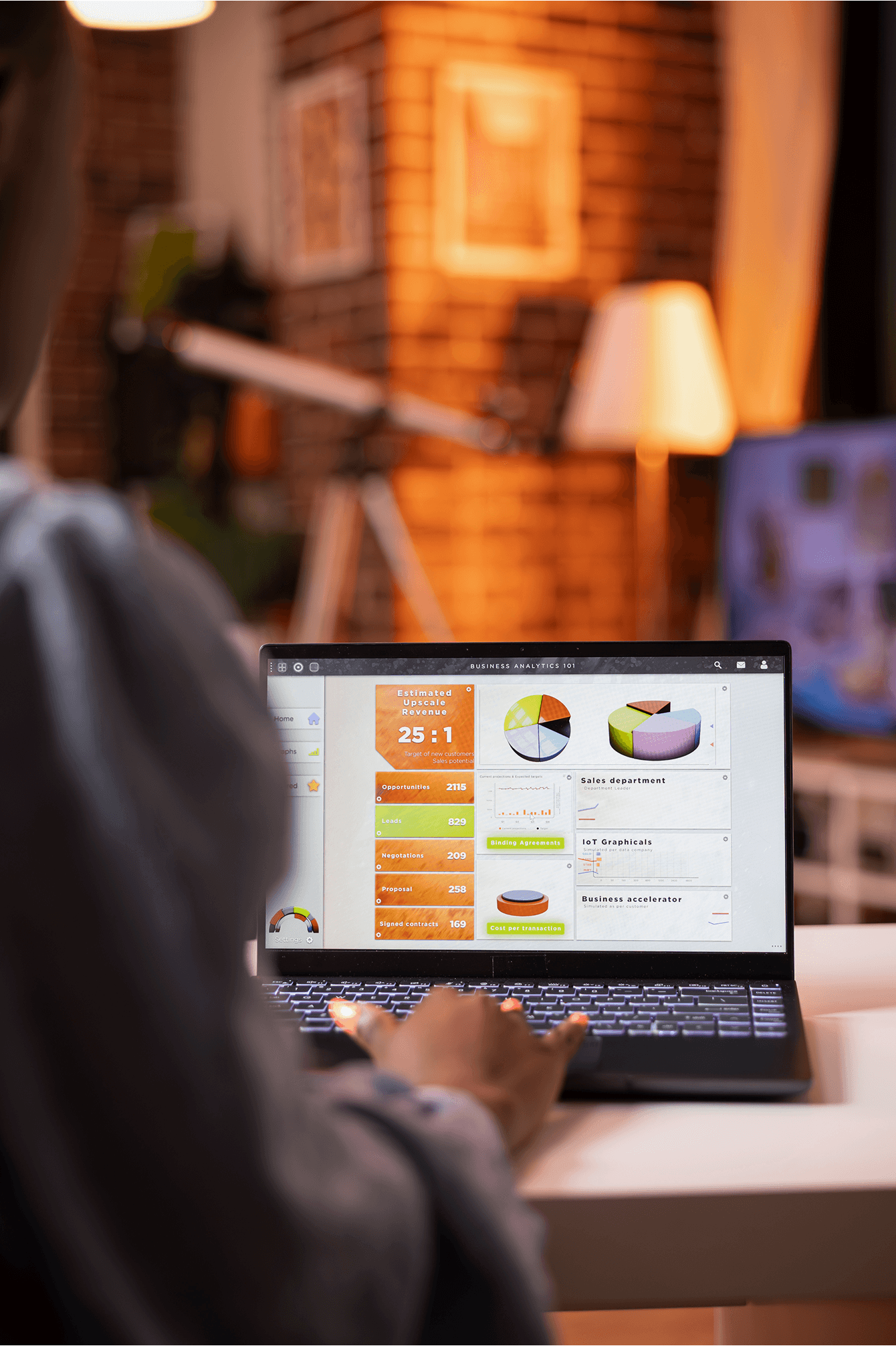 Over-the-shoulder view of a person working on a laptop at night, displaying a colorful "Business Analytics 101" dashboard with pie charts, sales metrics, and an "Estimated Upscale Revenue" ratio of 25:1 in a warm, dimly lit home office.