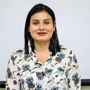 A woman with dark hair and a floral blouse smiles against a plain background.