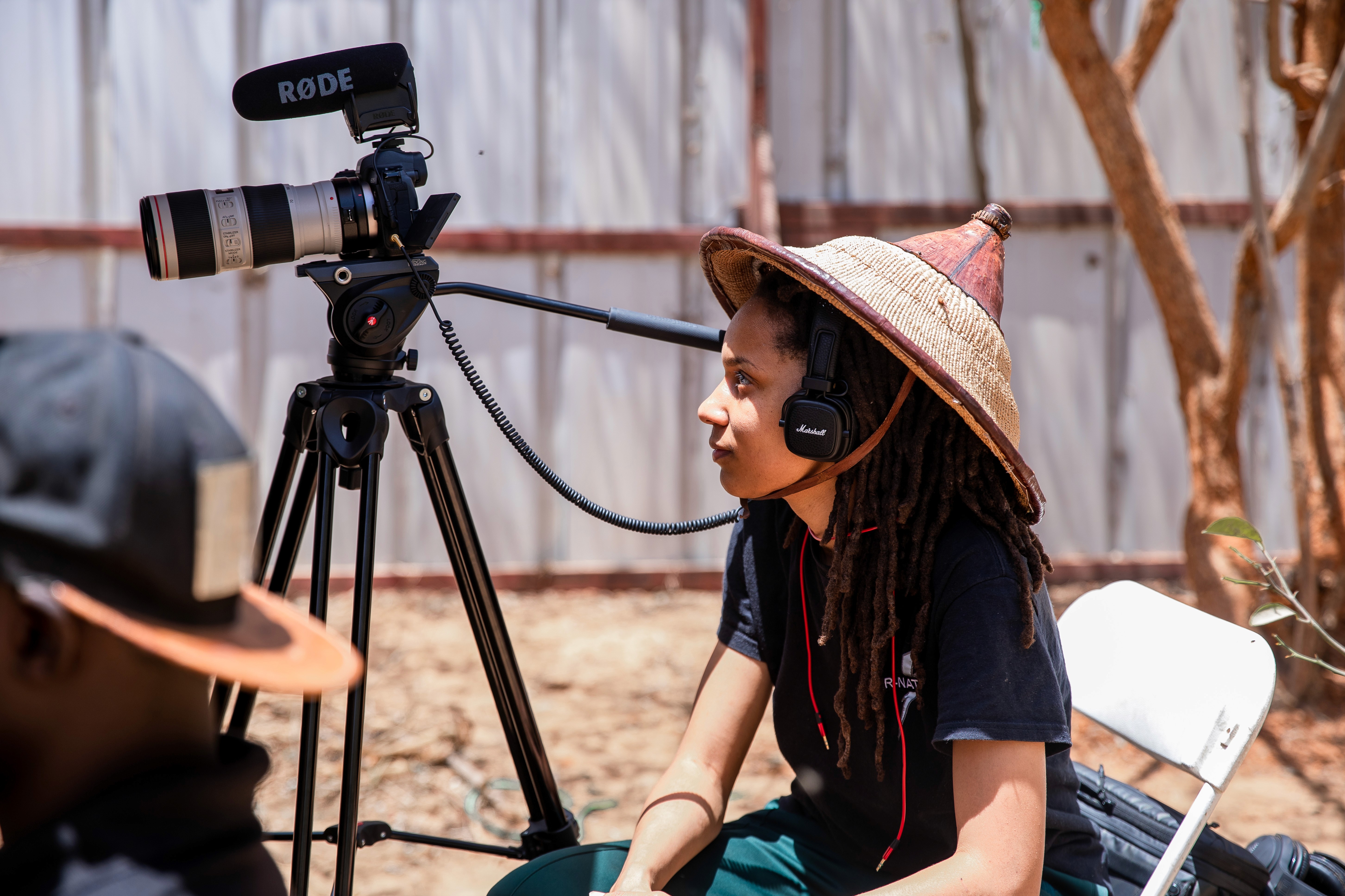 A side-profile of a young person with dark, wavy hair, wearing silver sports sunglasses and a black sweater, sitting in front of a blurred teal couch.