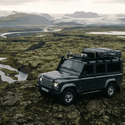 A grey Land Rover Defender parked on a rocky, mossy hill overlooking a wide river valley and mountains under a cloudy sky.