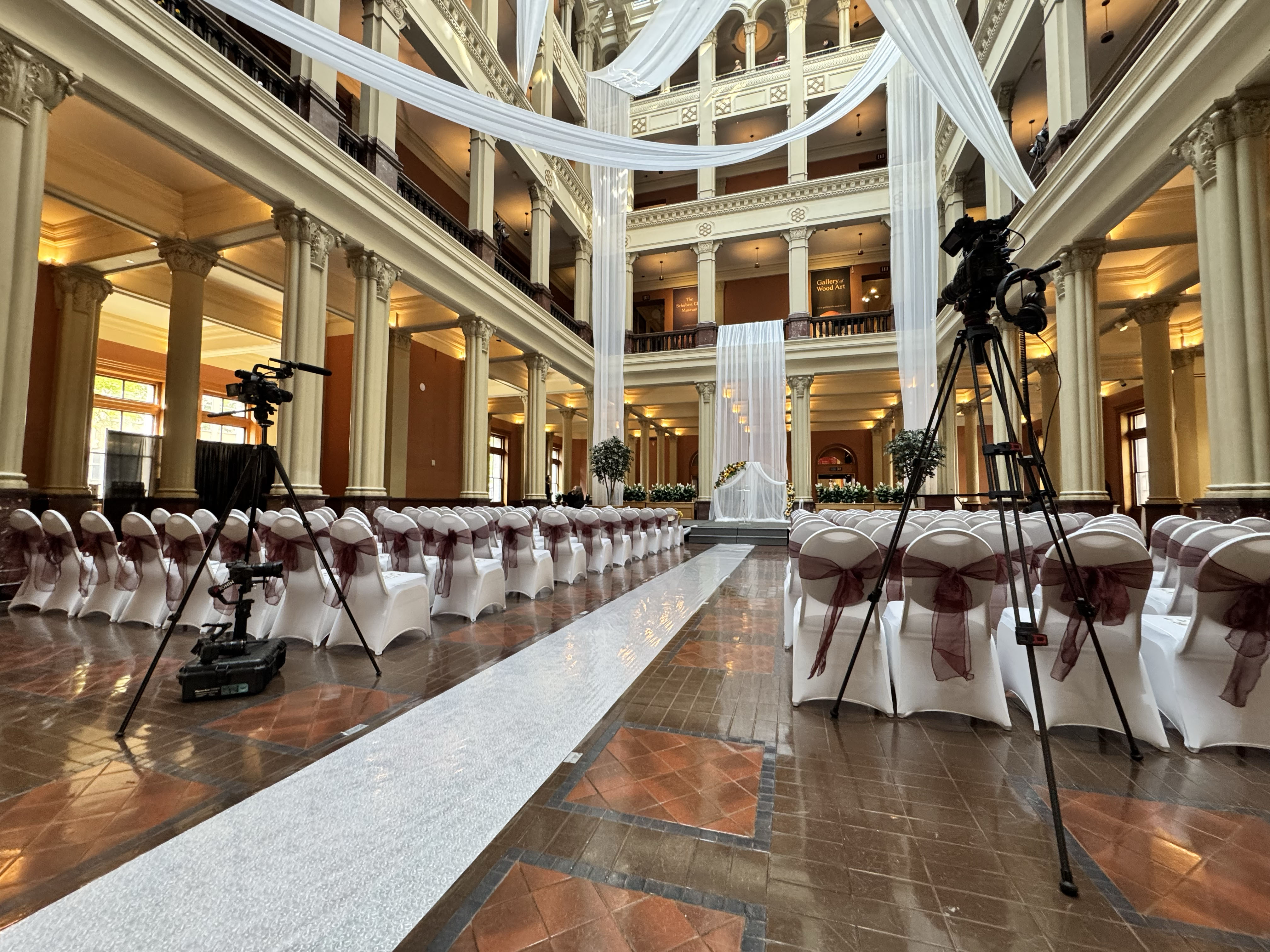 Professional video cameras on tripods set up inside a large, grand event hall with rows of white banquet chairs