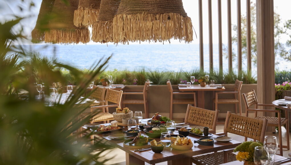 Ein gedeckter Tisch in einem Restaurant am Meer mit verschiedenen mediterranen Speisen unter rustikalen, geflochtenen Lampenschirmen mit Blick auf den Ozean.