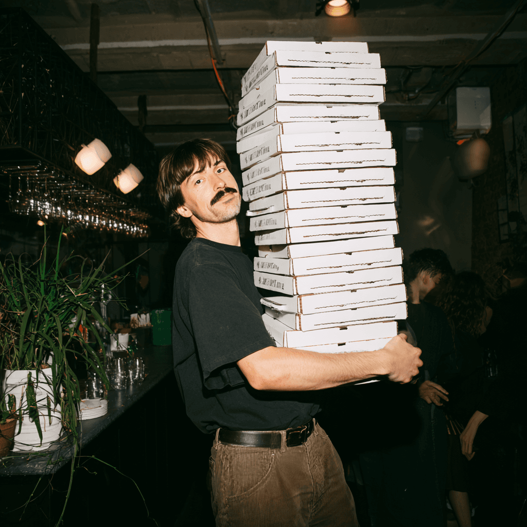 Person with a mustache wearing a black t-shirt carrying a tall stack of pizza boxes inside a dimly lit bar or restaurant.
