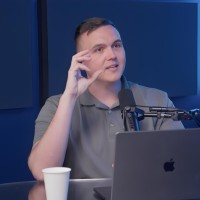 A smiling person with short, dark hair and a light-colored shirt against a dark background.