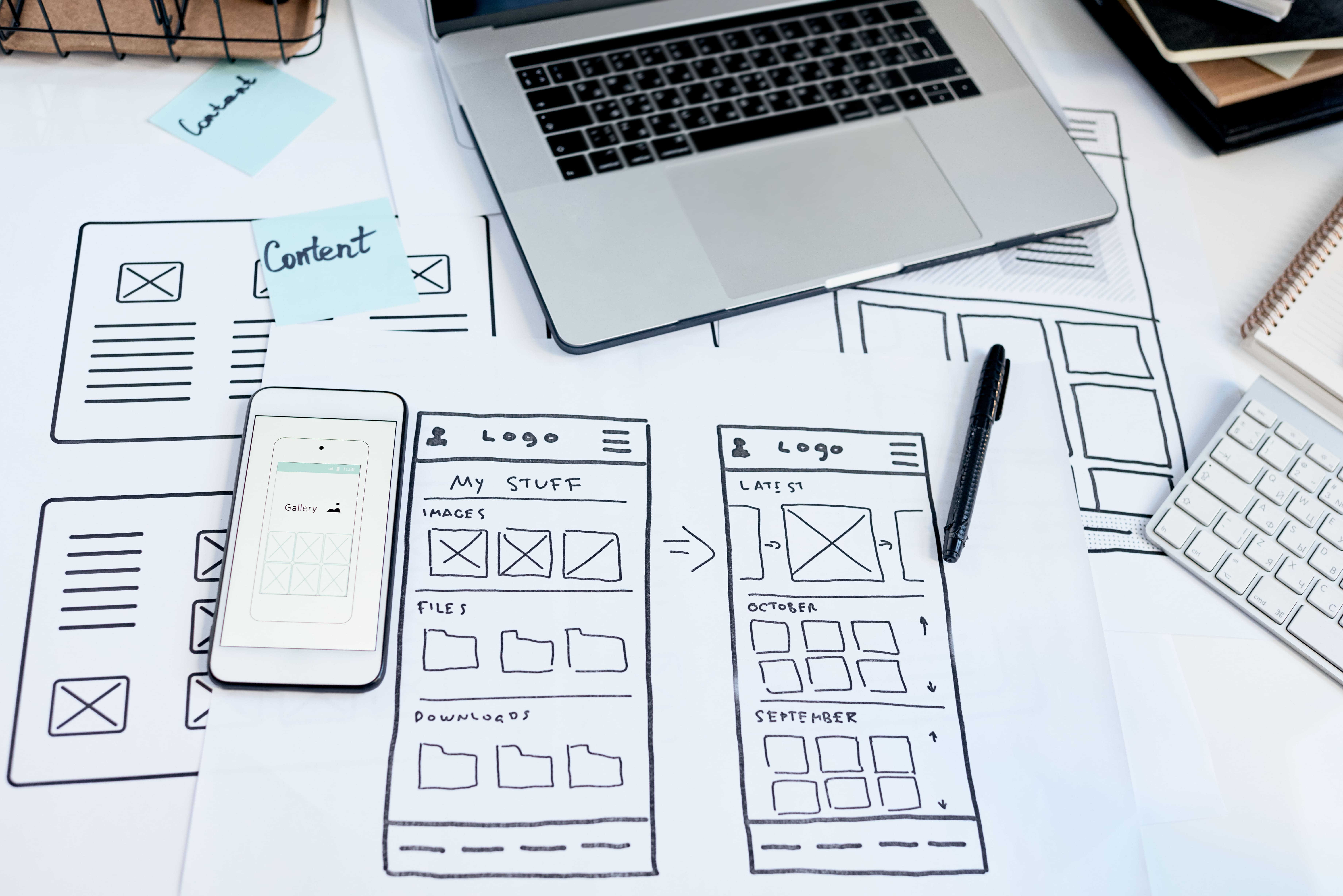 Flat lay of website wireframes and mobile UI sketches with laptop, smartphone, and keyboard – concept planning for web design and app development by a creative agency.