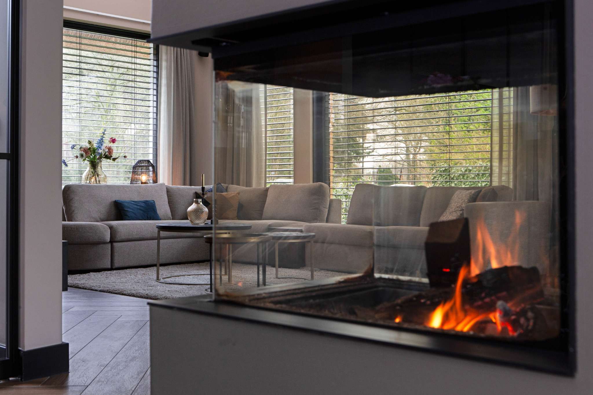 Woonkamer met hoekbank gezien door een moderne doorkijkhaard, in warme en tijdloze stijl.