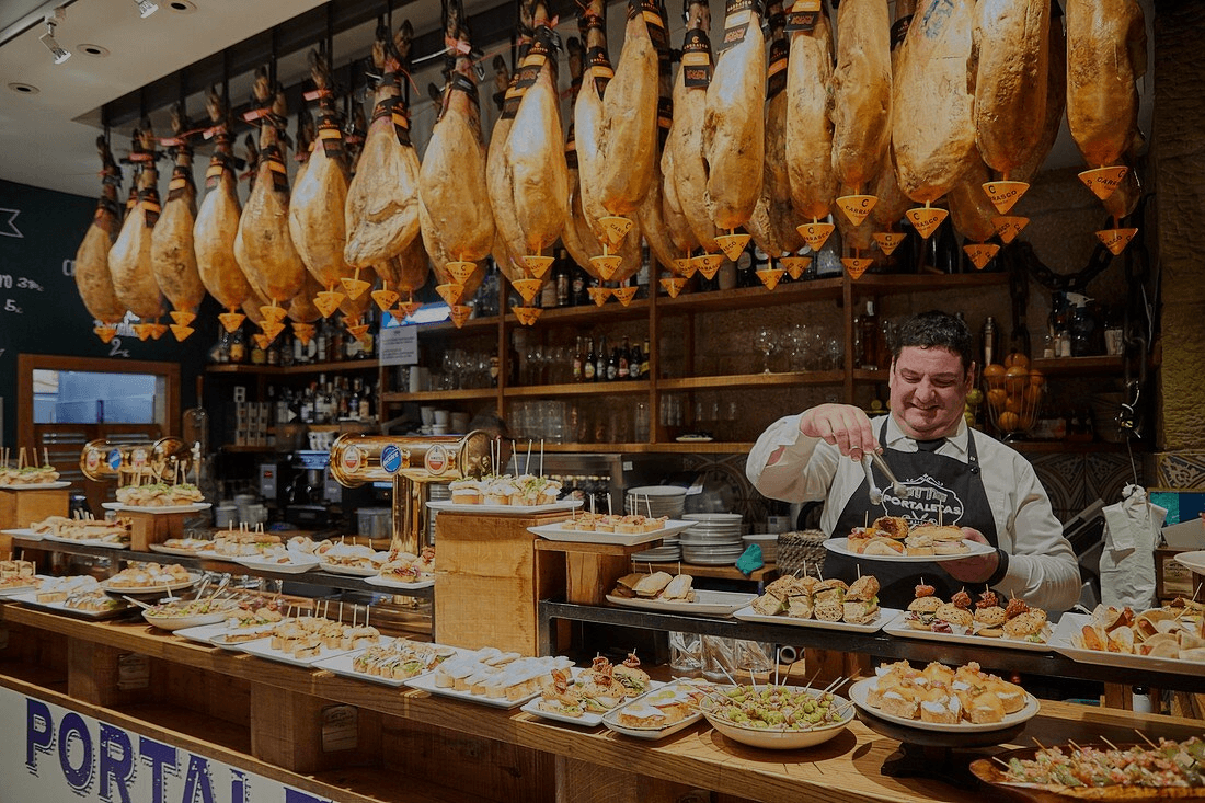 A bustling deli counter with hanging cured meats and a staff member preparing orders behind the counter.