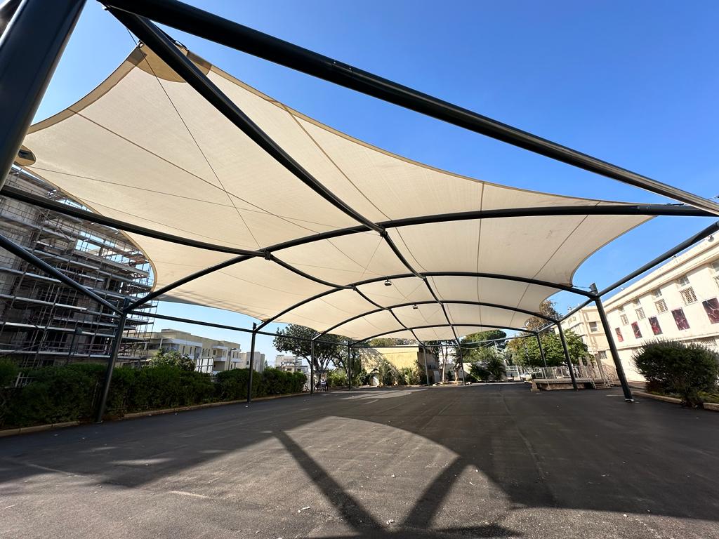 Large beige tensioned sunshades over a paved area with buildings and trees in the background..
