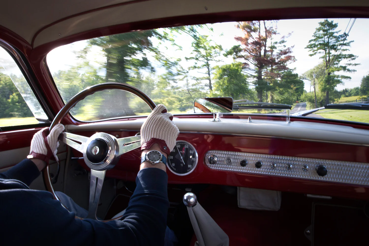 Driver wearing vintage-style gloves and a watch, steering a classic red car through a scenic, tree-lined road.