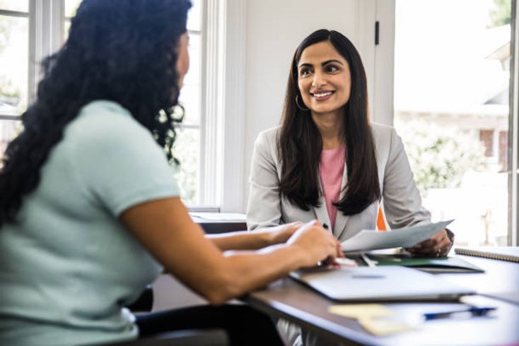 Two women sit at a table, smiling and discussing documents in a bright office. One wears a pink top and blazer, creating a positive atmosphere.
