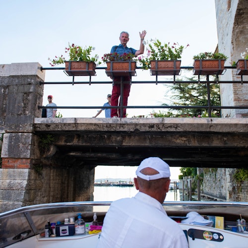 A man waves from a bridge adorned with flower boxes while another man steers a boat underneath. A tree and stone structure are visible.
