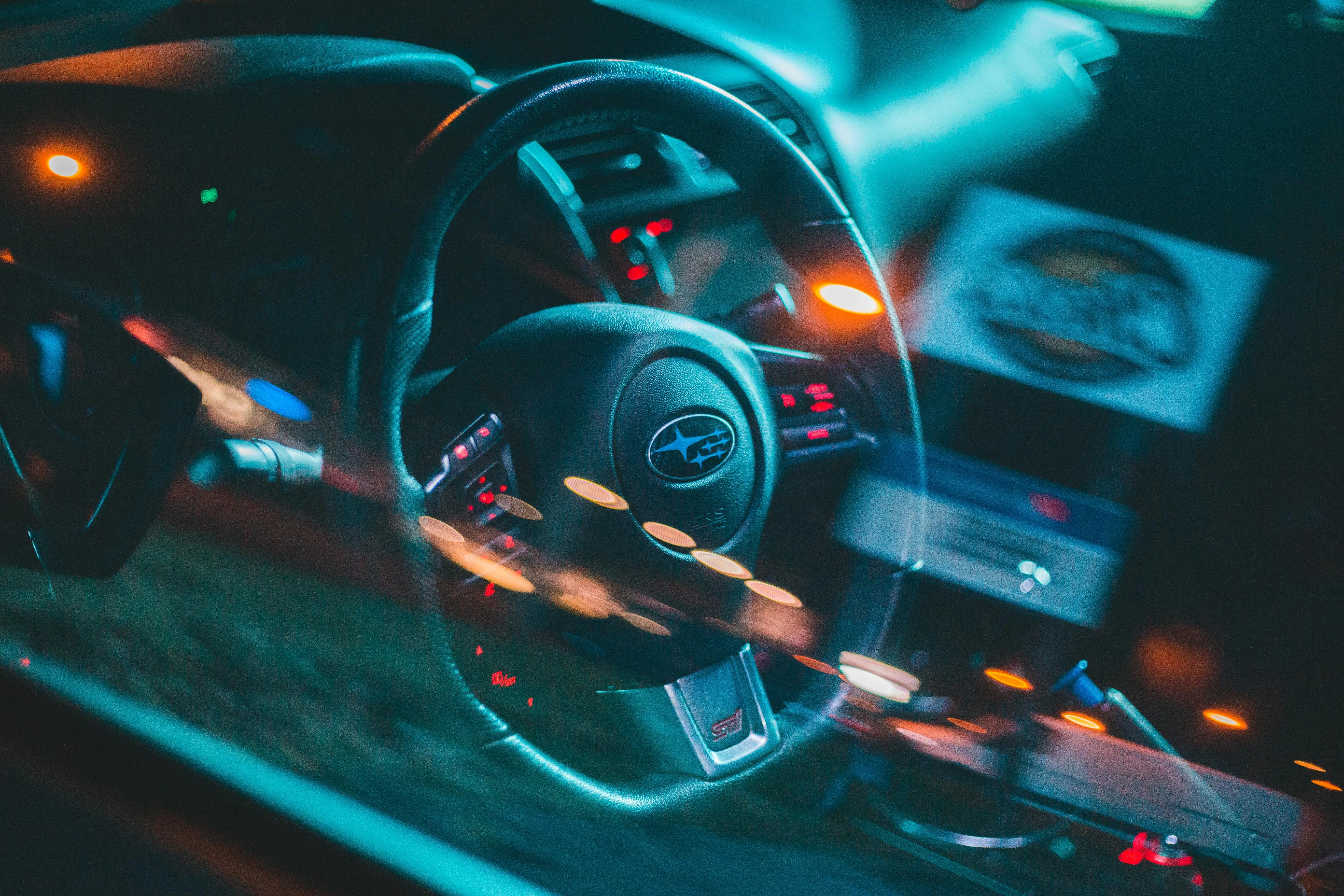 Vue intérieure d’une Subaru WRX, capturée de nuit à travers la vitre. Le volant siglé Subaru, les commandes rétro-éclairées en rouge et les reflets lumineux créent une ambiance dynamique et technique. L’éclairage bleu et les flous de mouvement renforcent l’atmosphère nocturne et sportive de la berline japonaise.