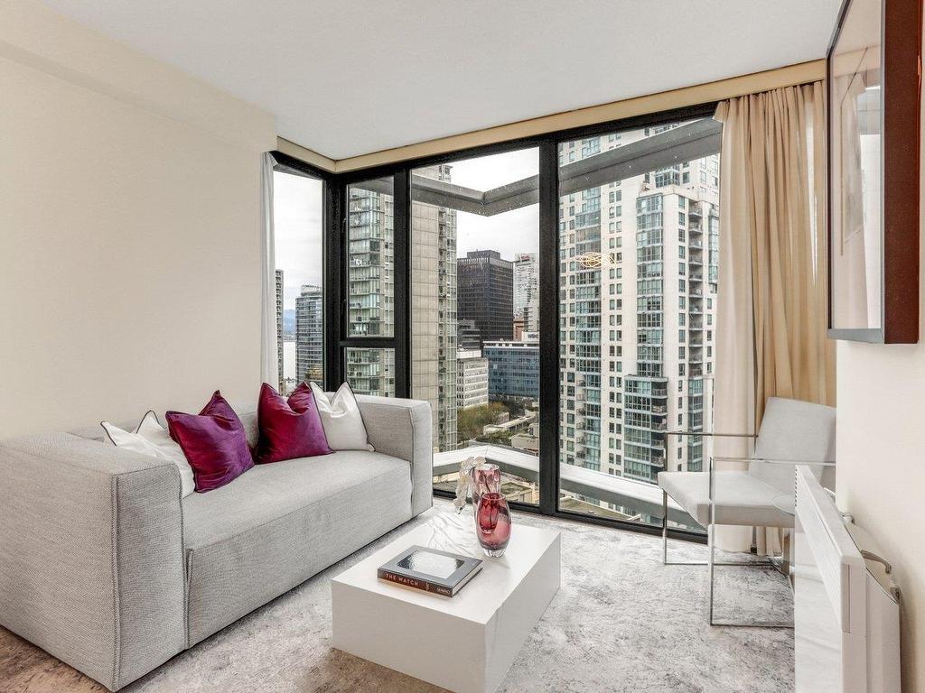 Modern living room with a gray sofa, purple cushions, and large windows overlooking a cityscape. Bright and airy space.