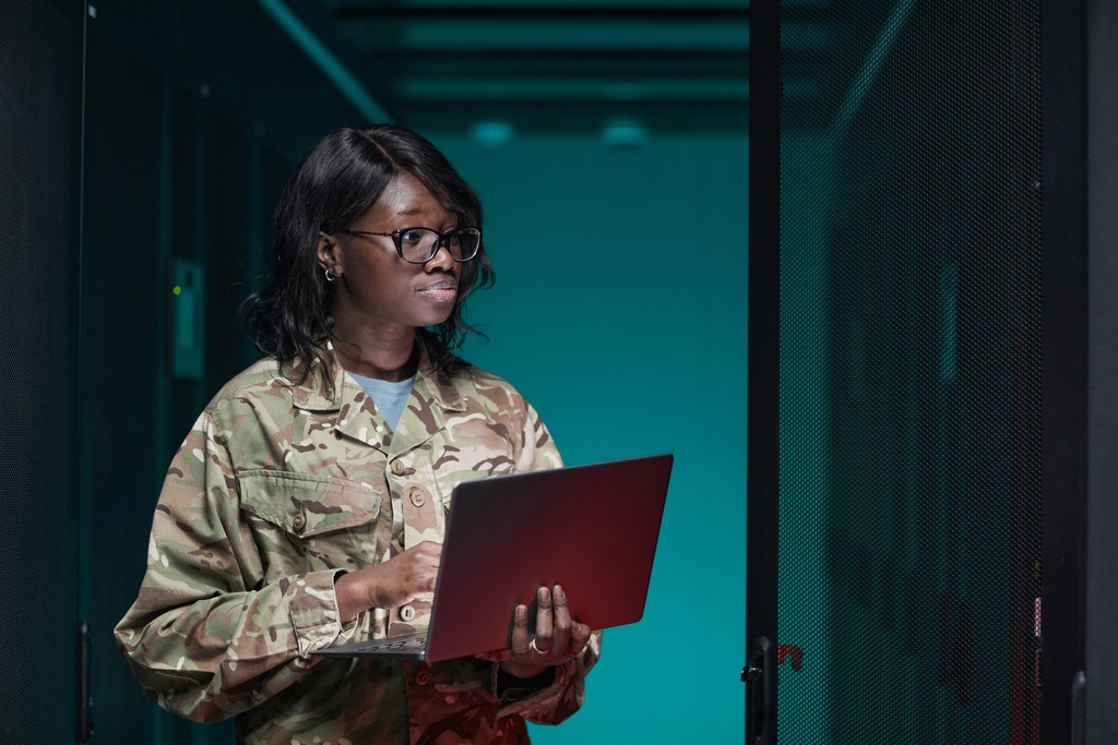 A military officer in camouflage attire works on a laptop inside a server room, illustrating the integration of technology and cybersecurity in modern military operations.