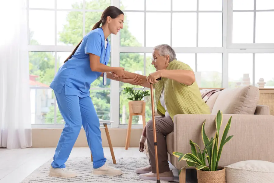 In-home caregiver in blue scrubs helping an elderly woman with a cane stand up from a sofa in a sunlit living room.