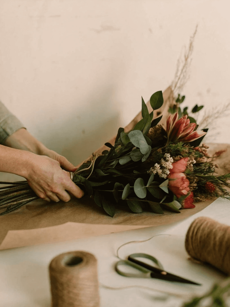 Hands arranging a bouquet of green foliage and pink-orange flowers.