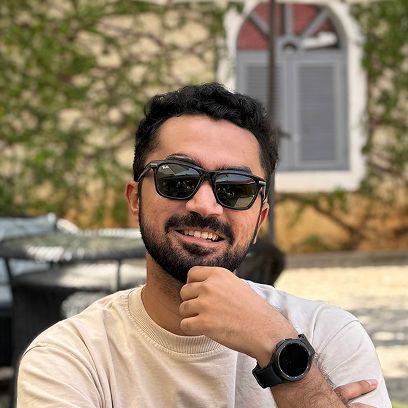 A man with a beard and sunglasses smiles, resting his chin on his hand, with greenery in the background.