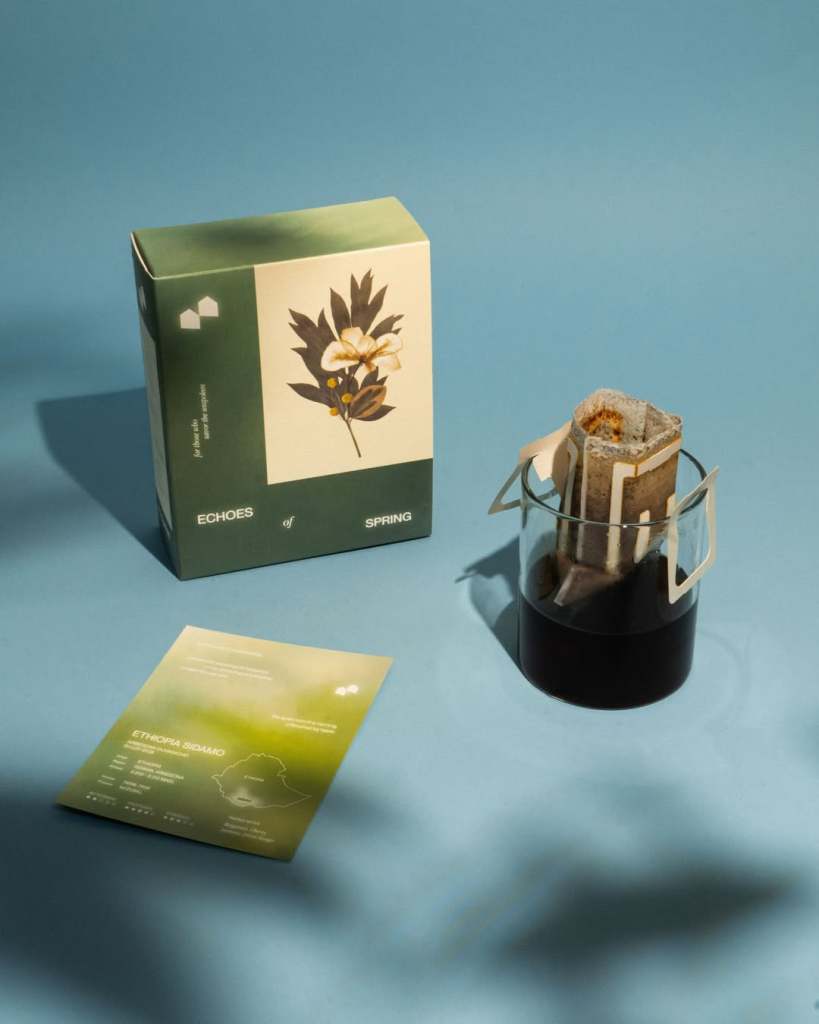 A box labeled 'ECHOES of SPRING' is placed next to a glass containing brewed coffee, with coffee brewing packets inside the glass and an informational card about Ethiopia Sidamo coffee on a blue surface.