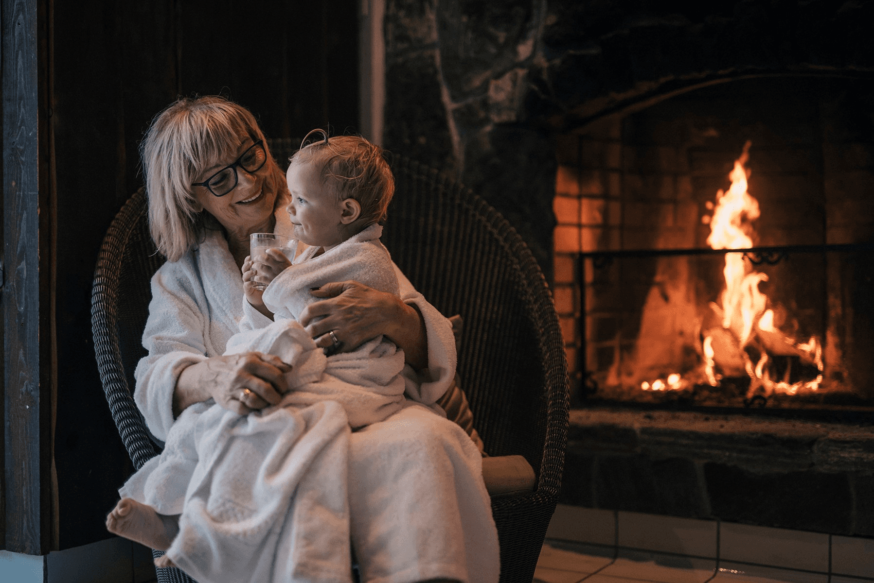 An elderly person wearing glasses, cozily seated in a wicker chair, embraces a toddler wrapped in a robe, in front of a crackling fireplace.