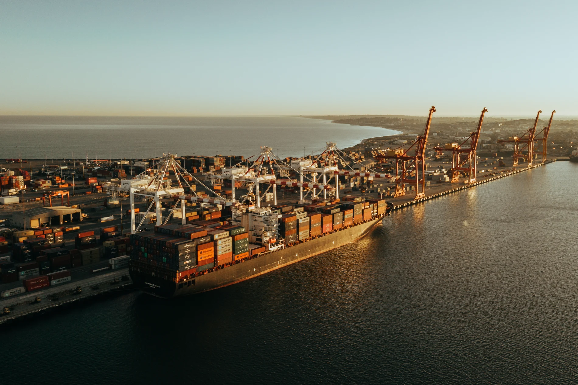 Aerial of a container ship berthed under gantry cranes at sunrise, coastline stretching beyond