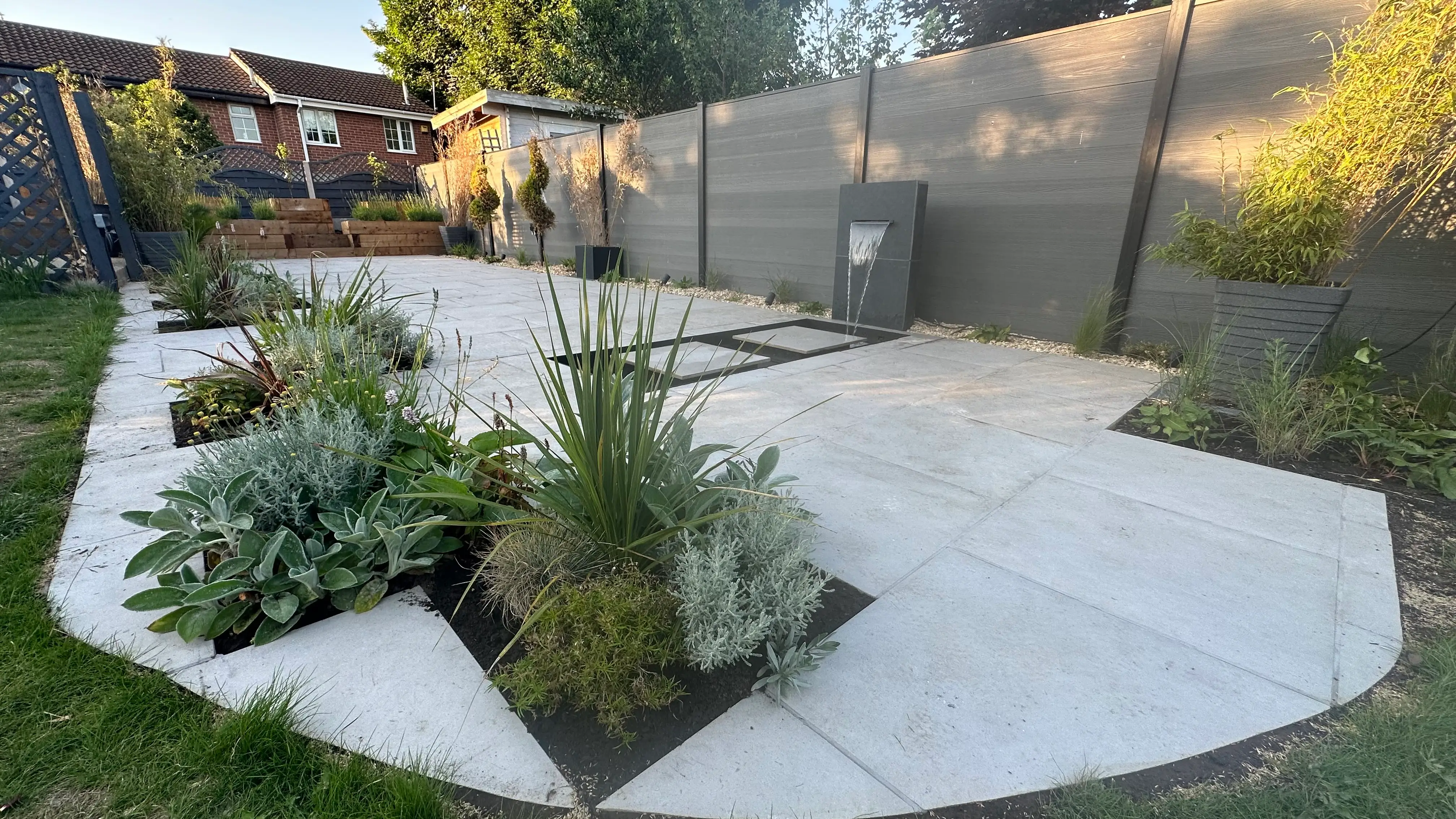 A modern garden featuring a geometric stone path and green plants, with a wooden fence in the background.