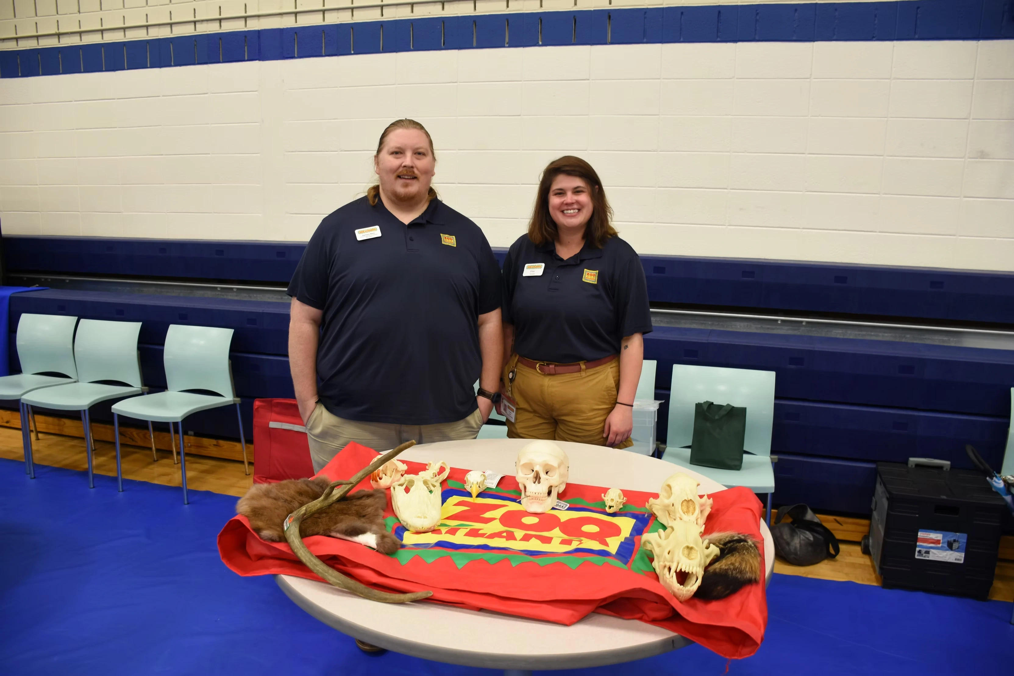 Two zoo staff members stand proudly behind a table displaying various animal skulls and pelts. The zoo's logo is visible.