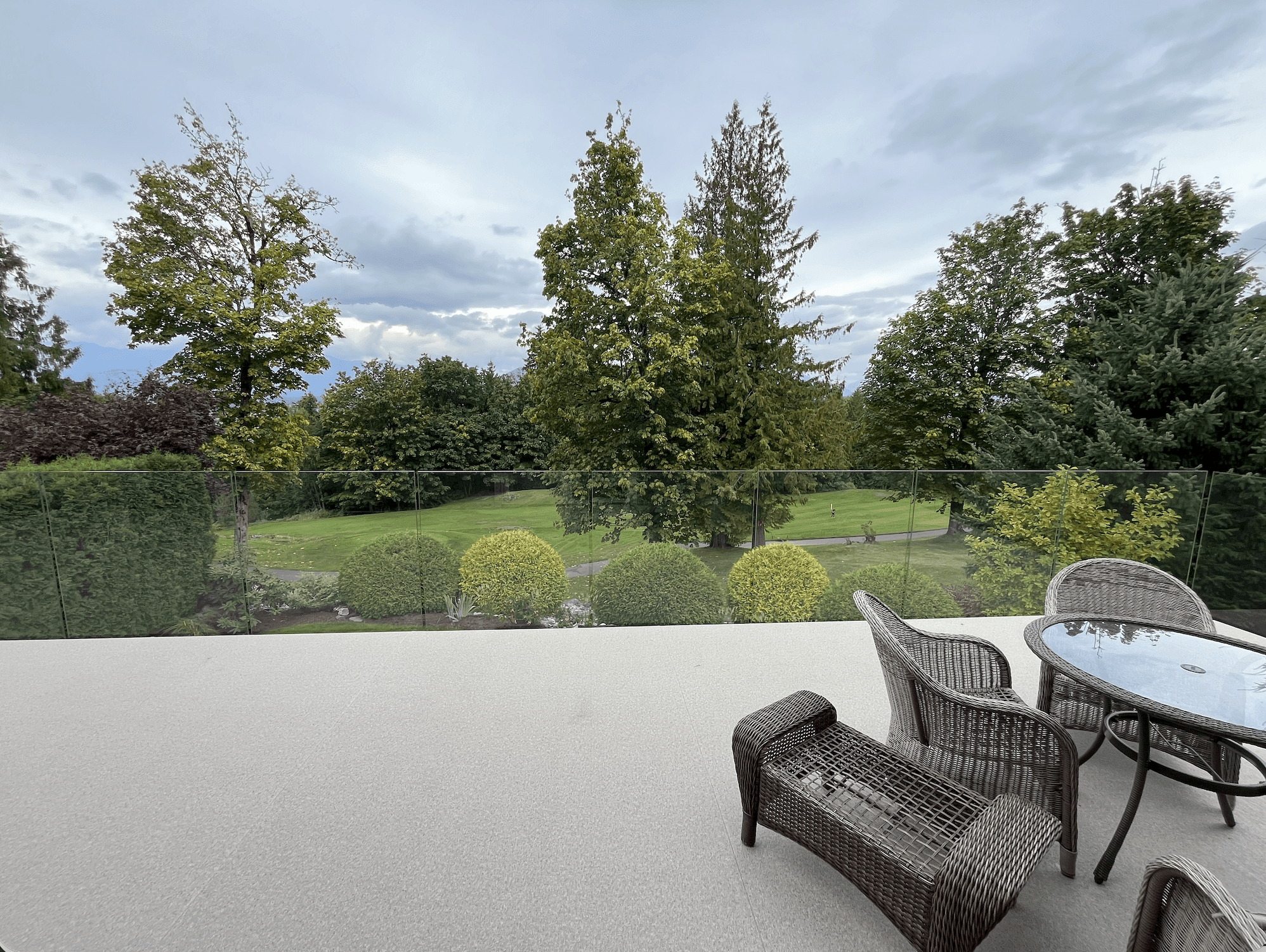 Outdoor patio with wicker furniture overlooking a landscaped backyard with manicured shrubs, trees, and a glass railing
