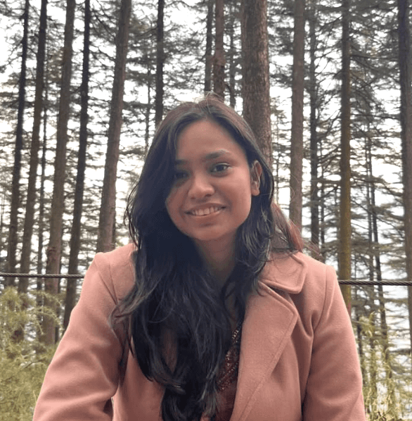 Person wearing a blue puffer jacket and red backpack standing in front of a light blue building on a city street.