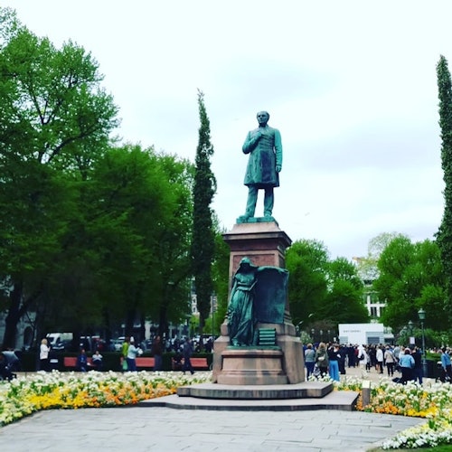 Χάλκινο άγαλμα ενός άνδρα σε βάθρο σε ένα πάρκο, περικυκλωμένο από ανθισμένα λουλούδια και δέντρα, με ανθρώπους που περπατούν κοντά.