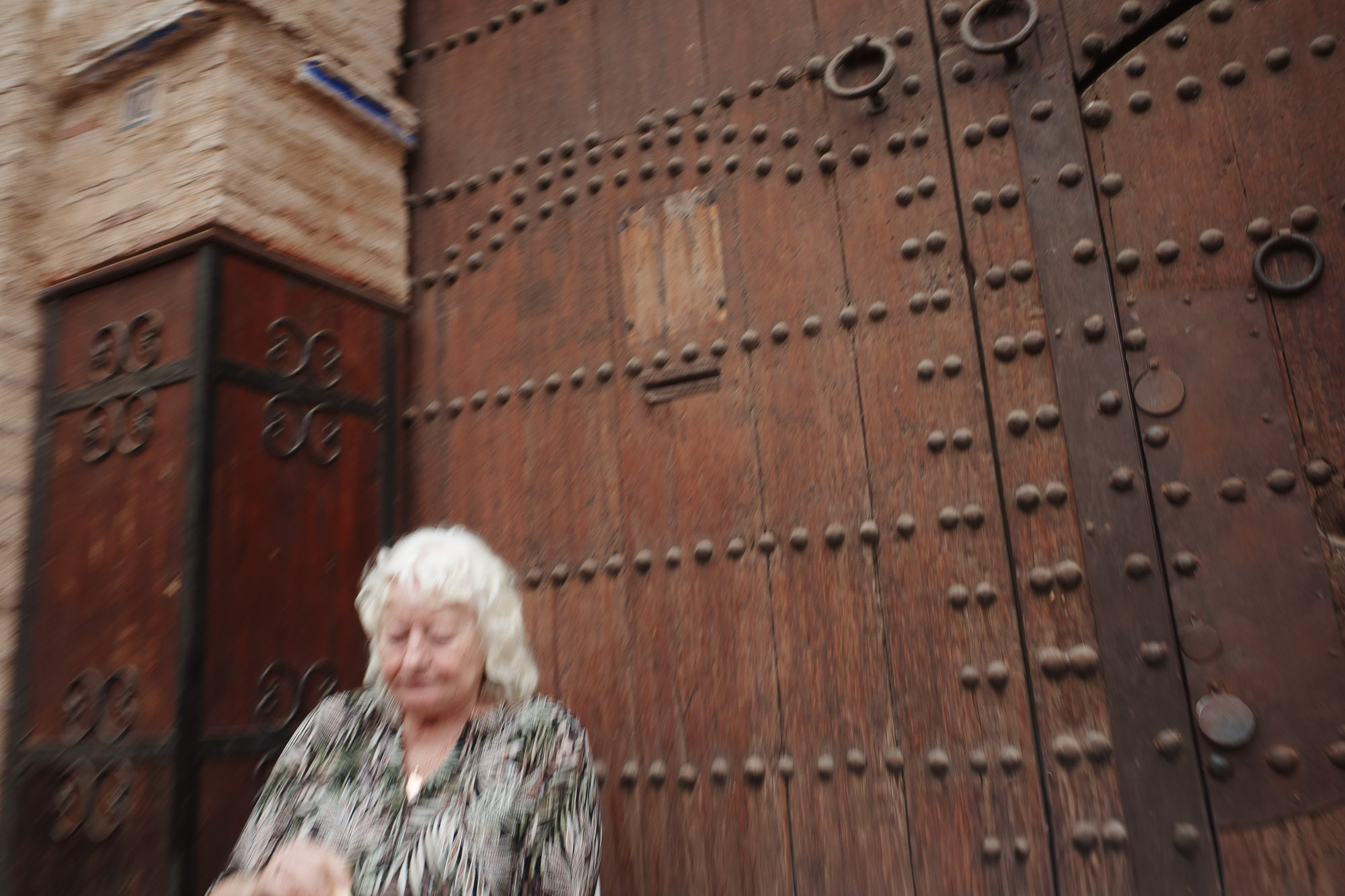 A person with white hair is sitting in front of an ornate, large wooden door with intricate metal detailing and a textured brick wall, creating a historic and rustic atmosphere. 