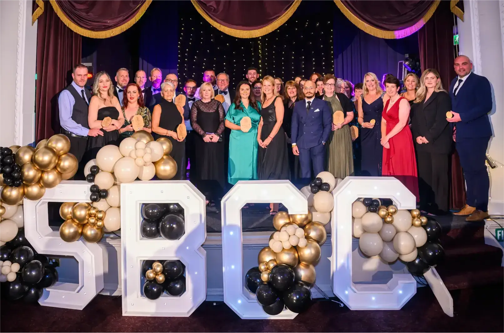 A group of people stands on a stage in formal attire, smiling. Large, illuminated letters "SBCC" are decorated with black, white, and gold balloons.