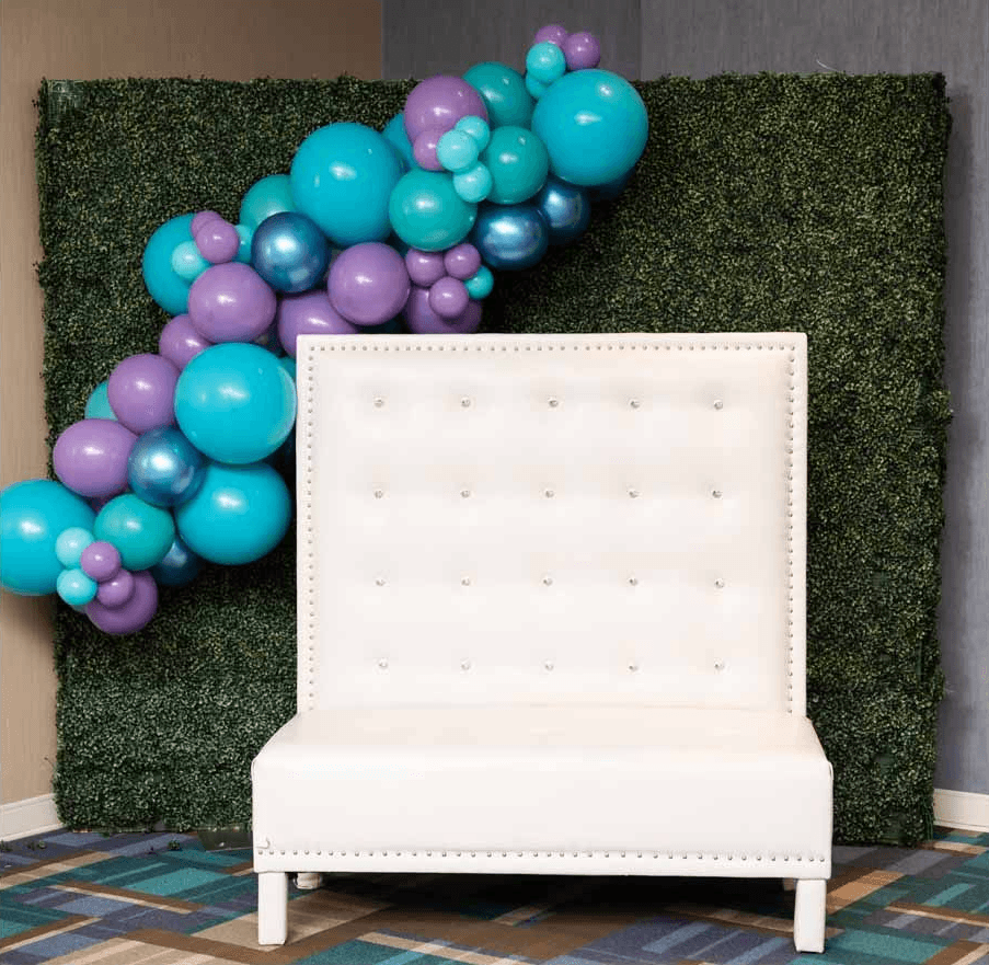 Blue and teal balloon garland framing a white tufted chair in front of a green boxwood backdrop created by Be Bold Balloons