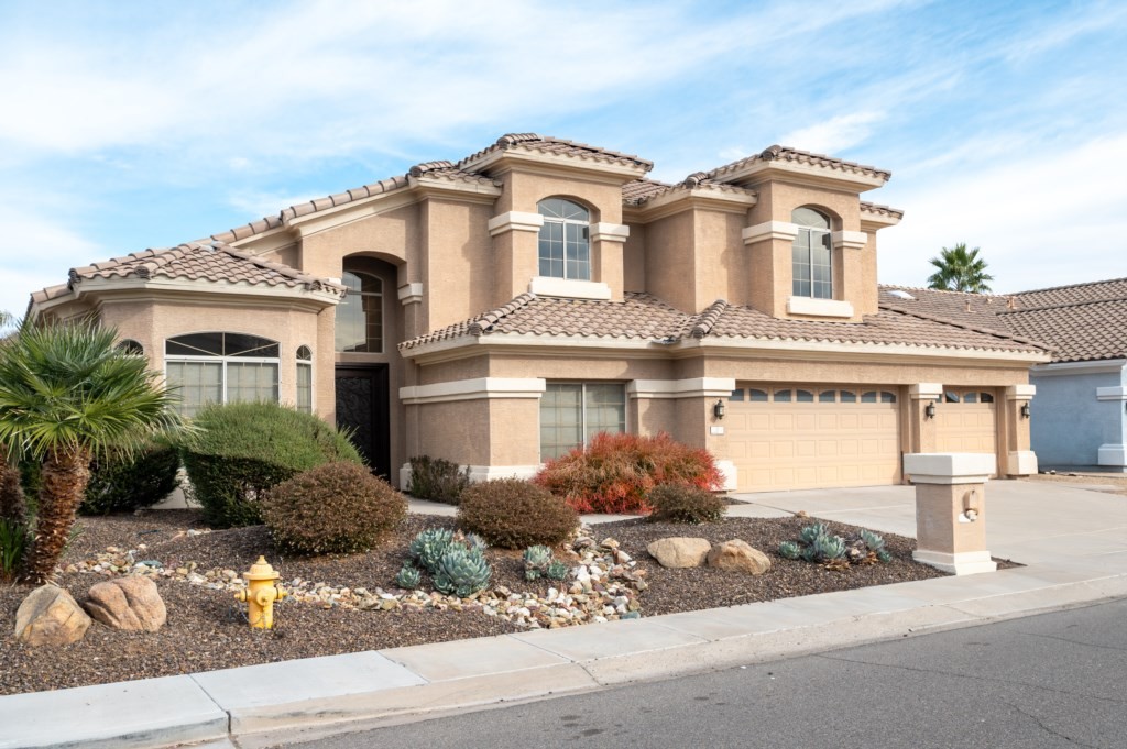 A spacious, modern two-story house with beige stucco exterior and tiled roof, featuring a landscaped yard with drought-resistant plants, representing an affordable luxury vacation rental house option.