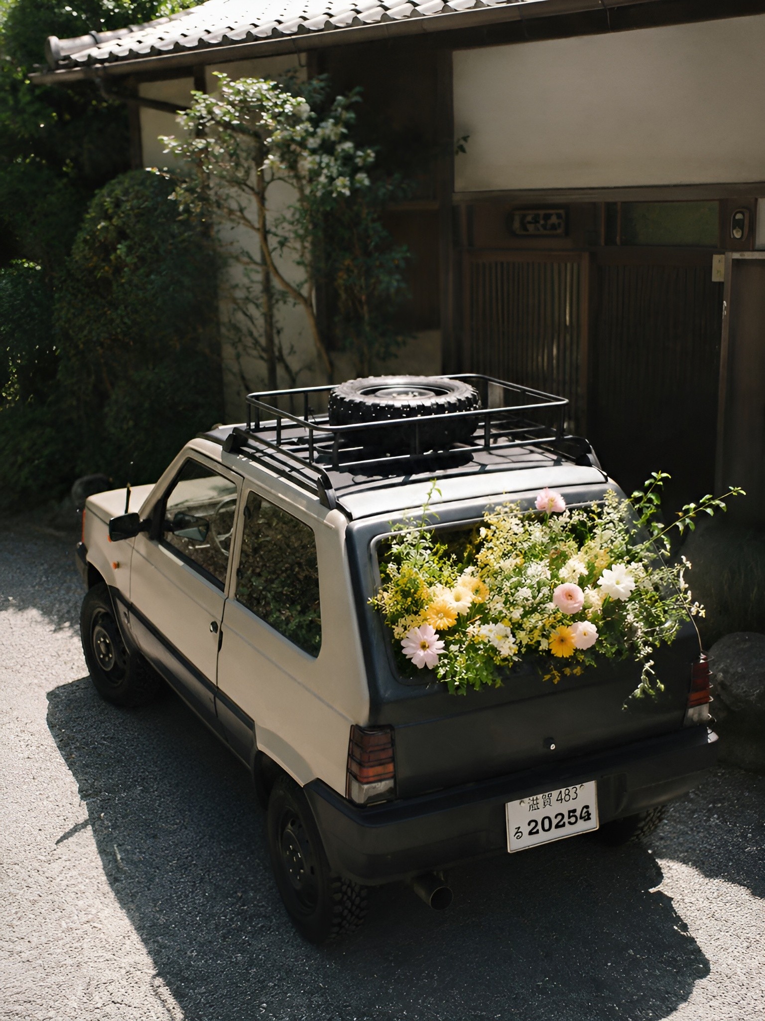 Car parked in a driveway with flowers growing from the open fuel tank, creating a surreal contrast within a quiet suburban setting.