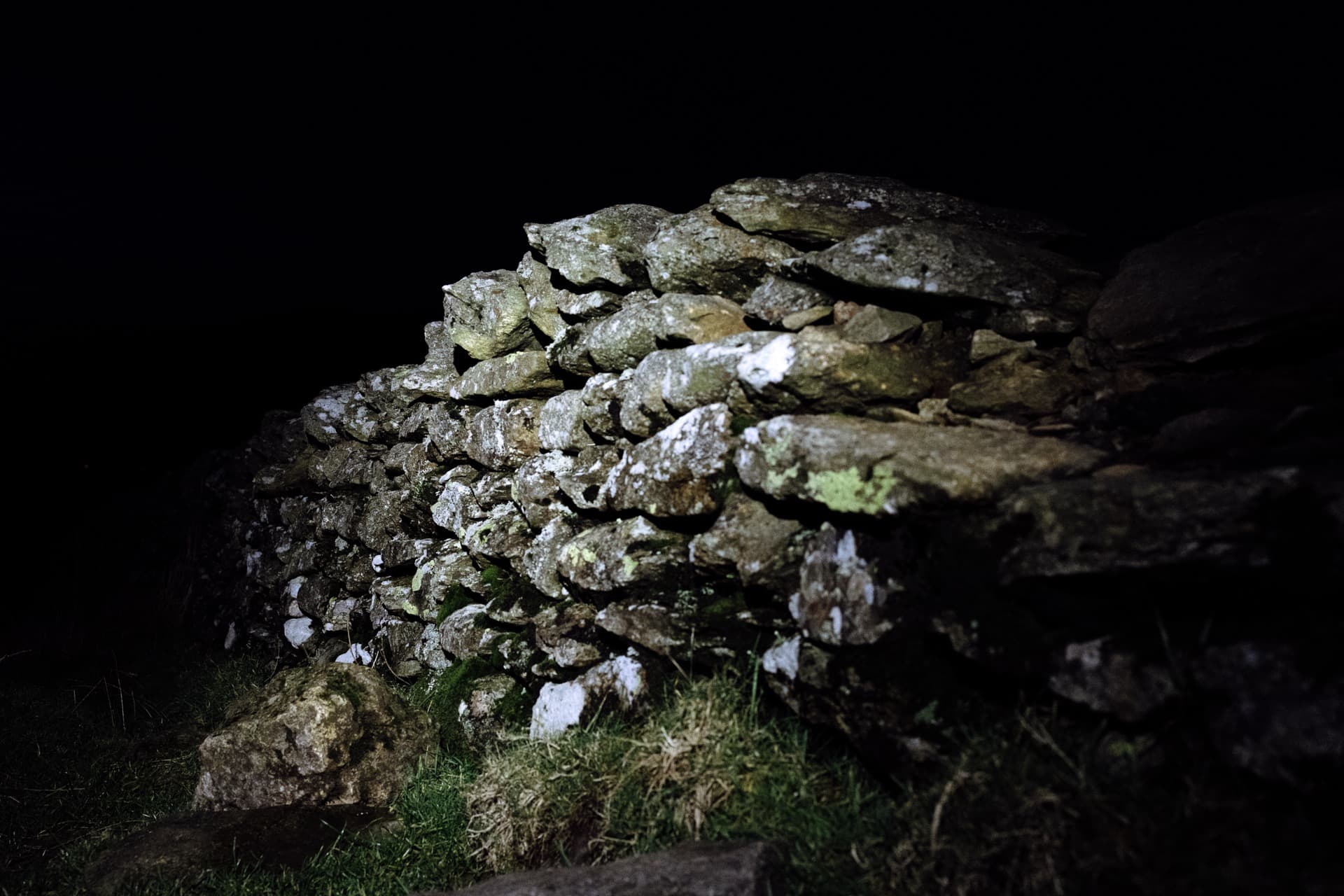 Dry stone wall section illuminated by headtorch against complete darkness, lichen and moss covering weathered slate stones, grass at base