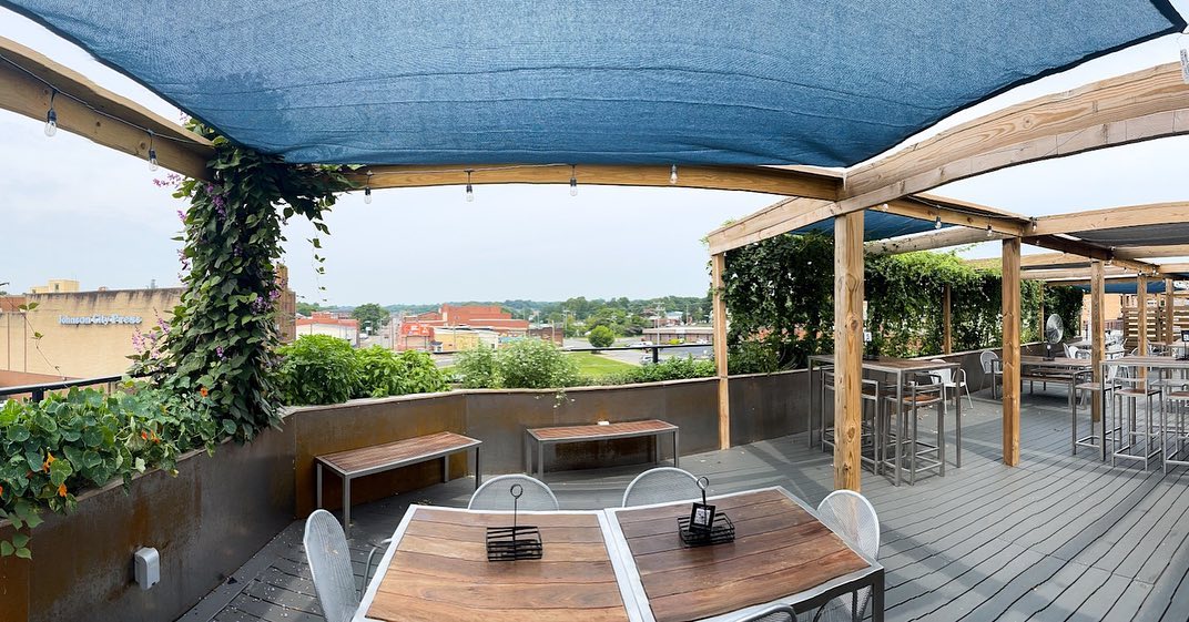 A serene outdoor dining area with wooden tables, shaded by a blue canopy, surrounded by green landscaping.