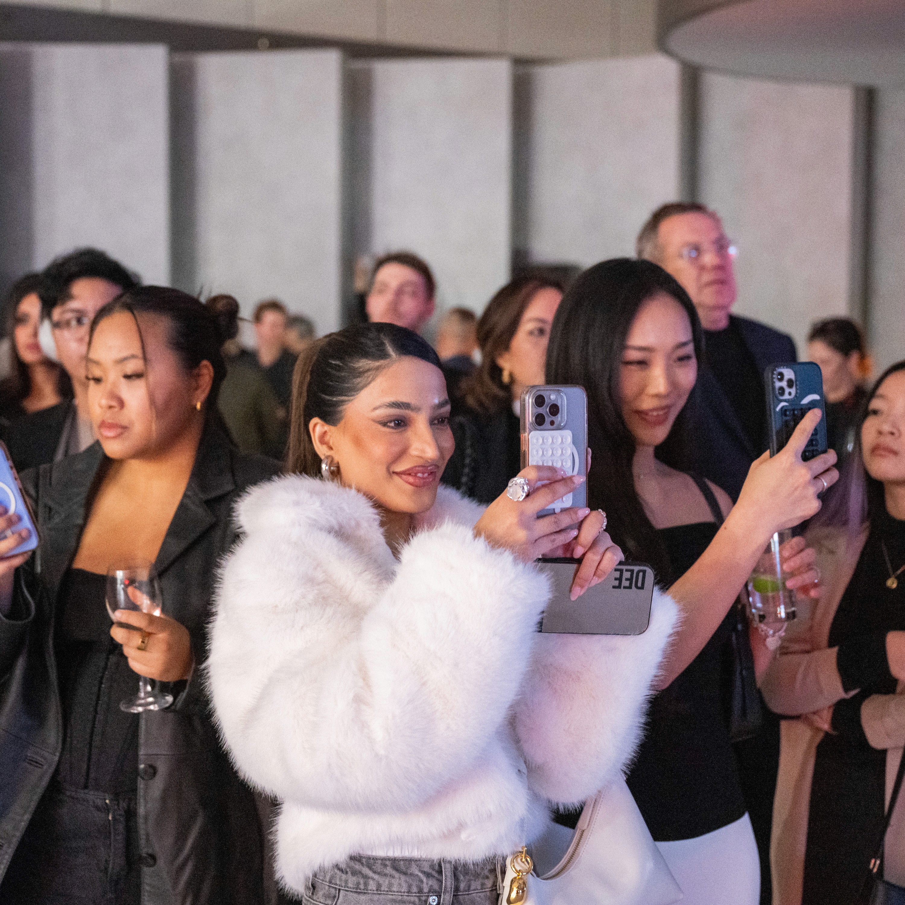 A group of people at the Audi showroom event, capturing moments on their phones. A woman in a white fur coat smiles, conveying excitement and engagement.