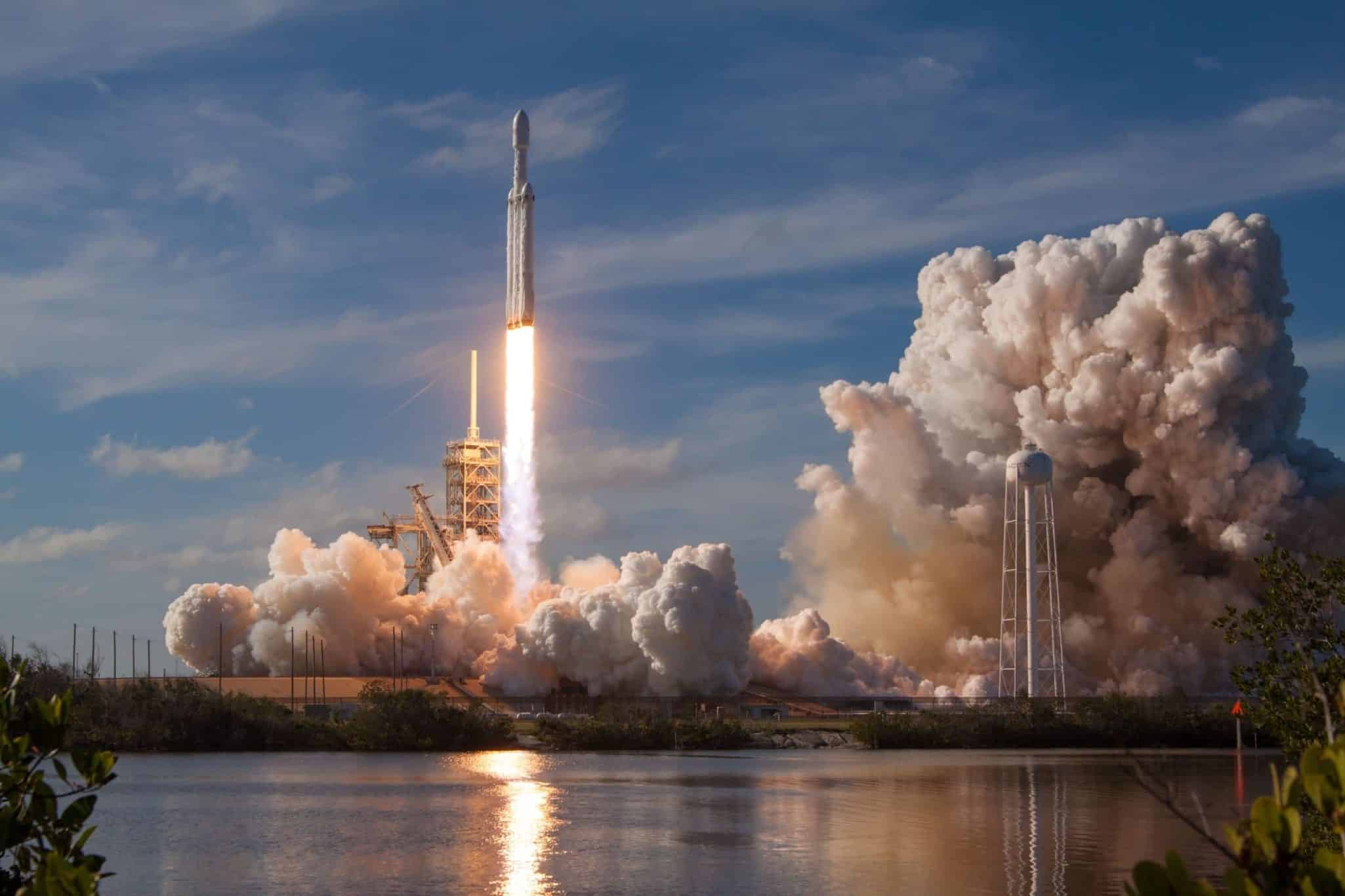 Rocket launch with vibrant plume of smoke and clear blue sky reflecting in water.