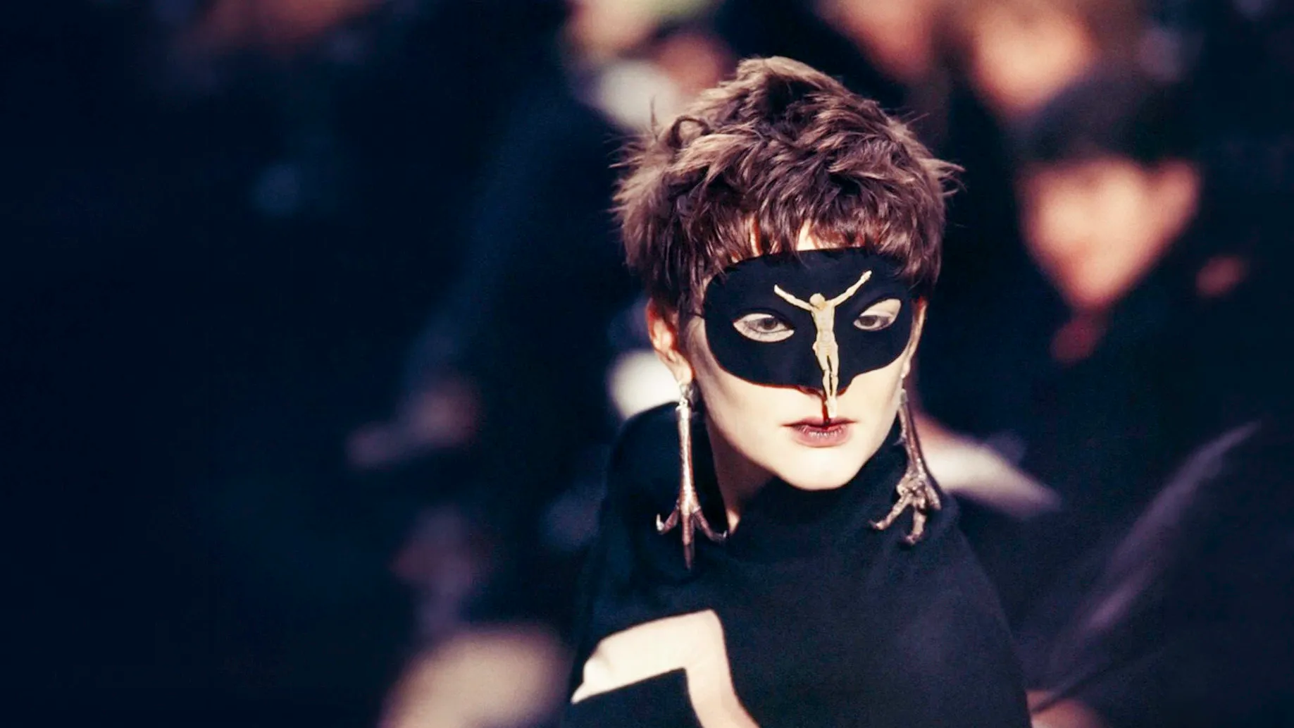Model on a dark runway wearing a black lace mask and a silver pheasant claw earring designed by Shaun Leane for the Alexander McQueen Dante collection.