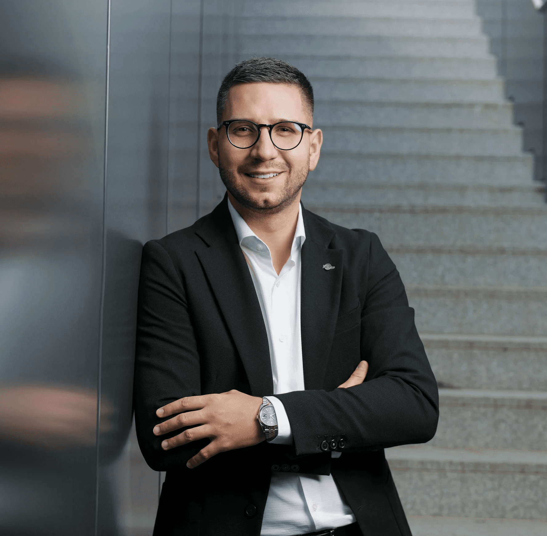 Muž s brýlemi a krýtkými vousy v bílé košili a černém saku opírající se o stěnu z ocelových panelů