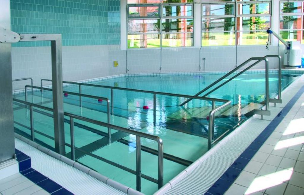 Indoor aquatic facility with turquoise water, metal safety railings, steps, and observation windows for therapy sessions.