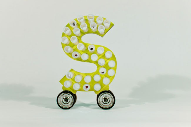 The image shows a large, three-dimensional letter "S" constructed from numerous white bottle caps arranged in rows. The "S" is light green or lime green in color and has small dark markings or designs on its surface. The letter is mounted on four black wheels with silver rims and is positioned against a plain white background. The wheels are slightly angled, suggesting movement. The composition is simple and focused on the letter "S".