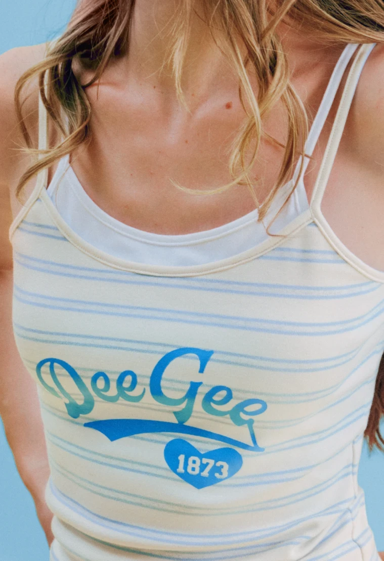 Close-up of a woman wearing a cream and blue striped tank top with a blue "Dee Gee 1873" heart logo, against a light blue background.