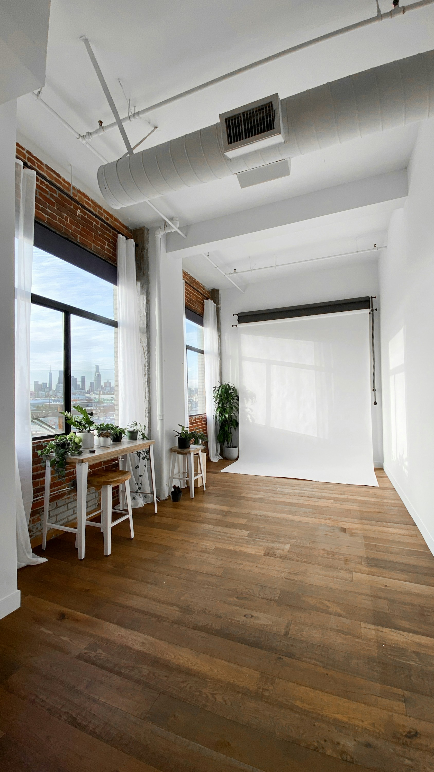 A bright and airy photography studio featuring exposed brick walls, large windows with sheer curtains, and a white backdrop roll for photoshoots. The wooden floor adds warmth to the space, and potted plants are placed around, adding a touch of greenery. The city skyline visible through the window suggests an urban location.