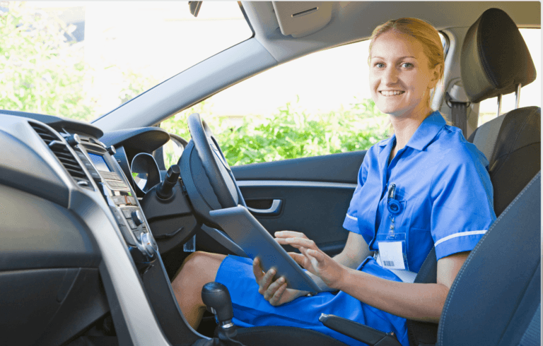 A female nurse in blue scrubs sitting in a car, smiling and holding a tablet or clipboard