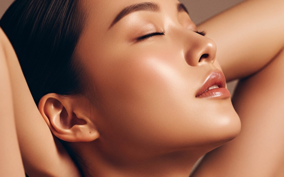 Close-up of a woman with her eyes closed, head tilted back, and arms raised. Her skin glows softly, conveying serenity and confidence.