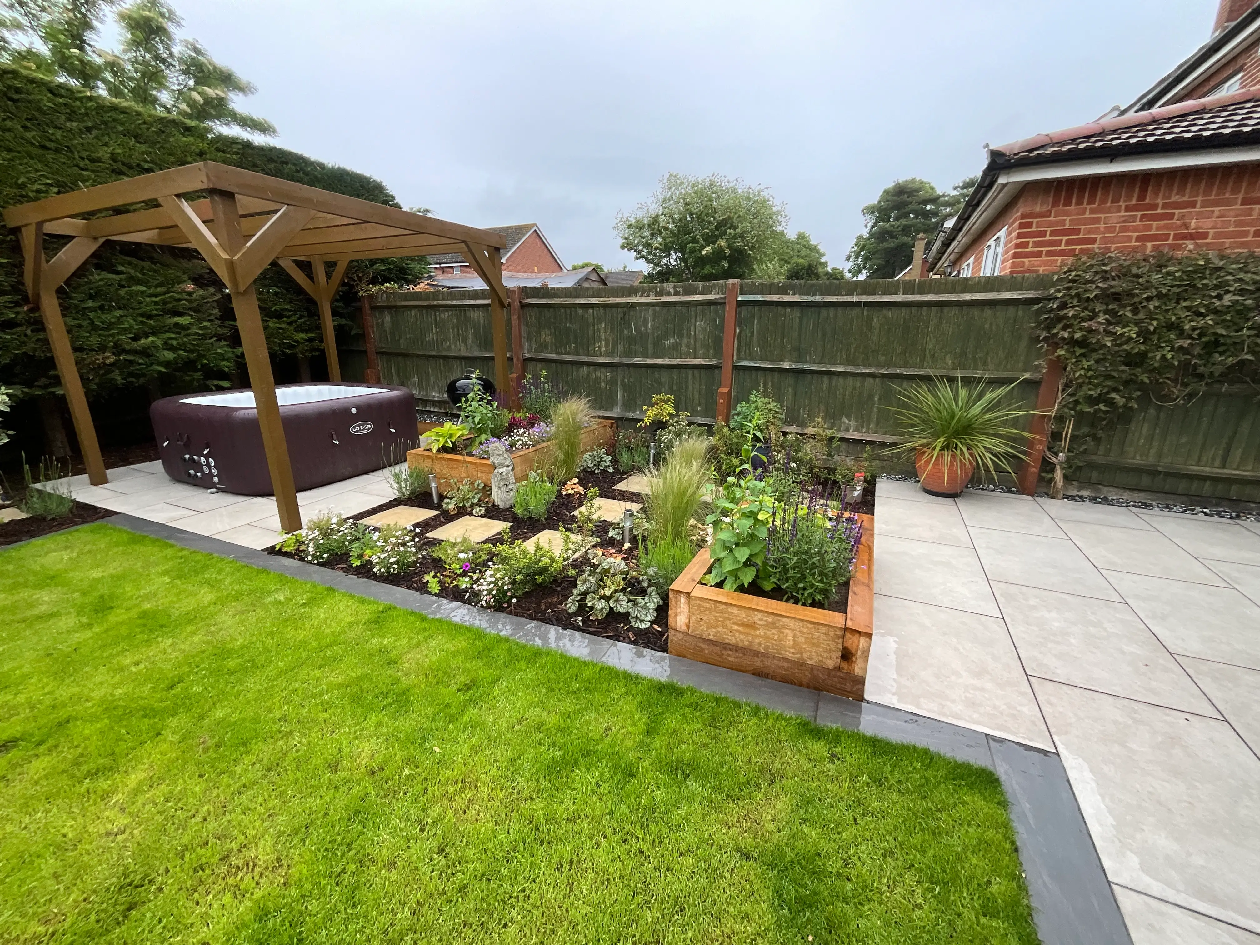 A landscaped garden featuring a hot tub, greenery, and stone pathways under a cloudy sky.