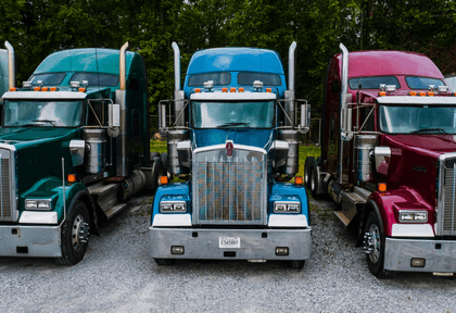 Three semi-trucks in green, blue, and red parked side by side on a gravel lot.