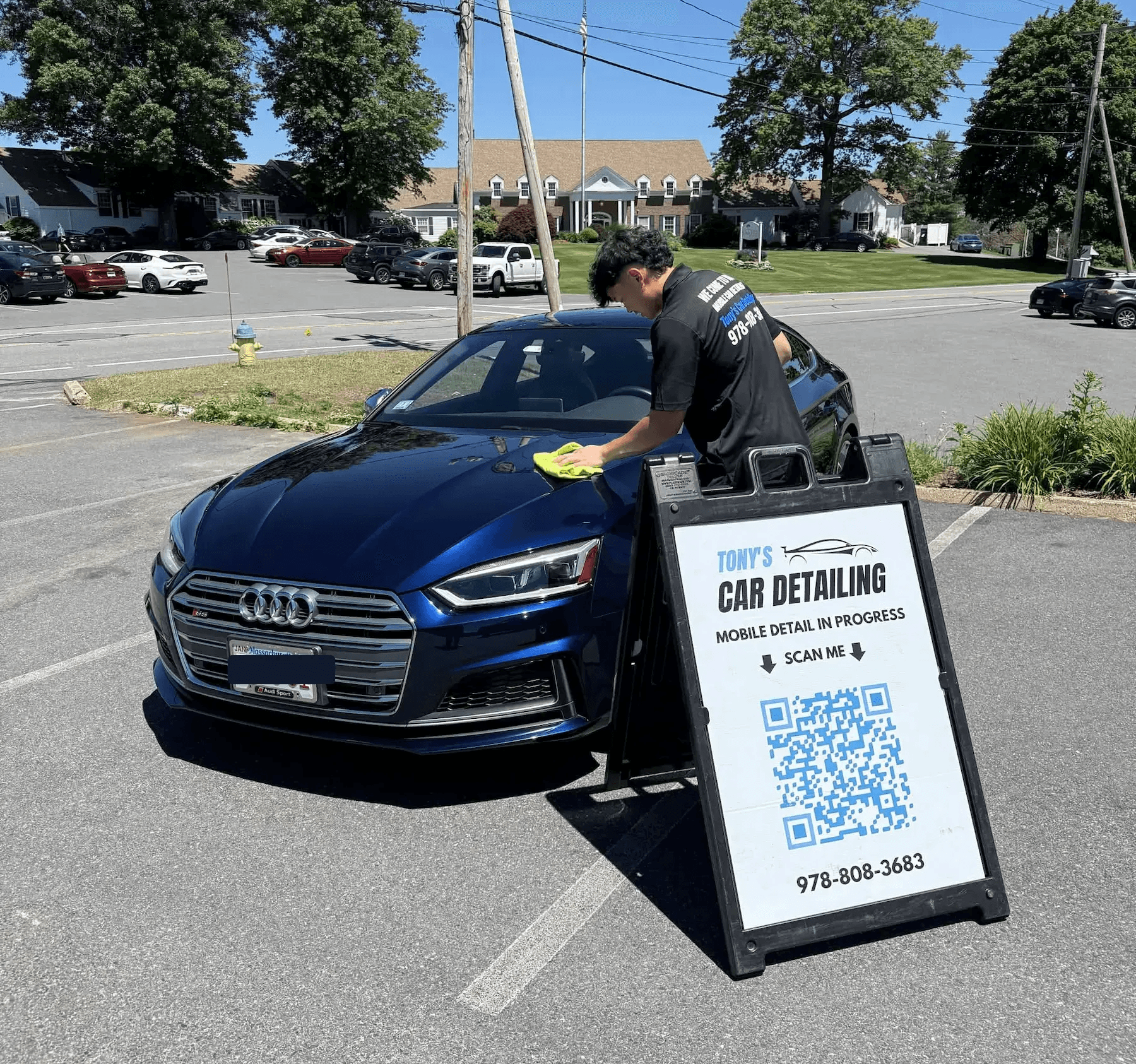 A mobile detailer wiping down the hood of a shiny, dark blue Audi sedan in a parking lot on a sunny day next to a "Tony's Car Detailing" A-frame sign with a QR code.